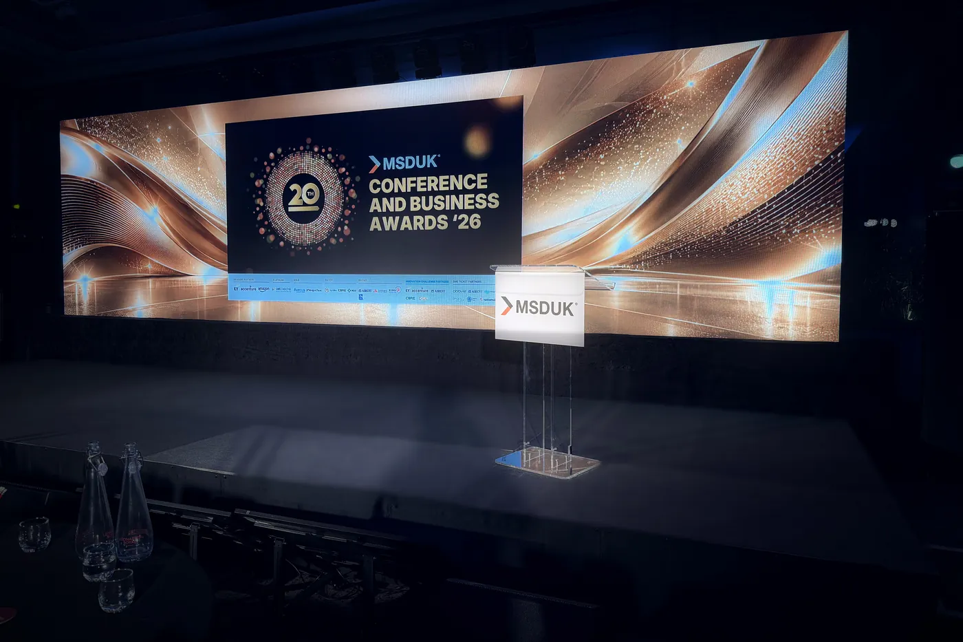 Stage for the MSDUK Conference and Business Awards 2026: a wide LED wall with the event branding and sponsor logos, a clear acrylic lectern and a carpeted stage