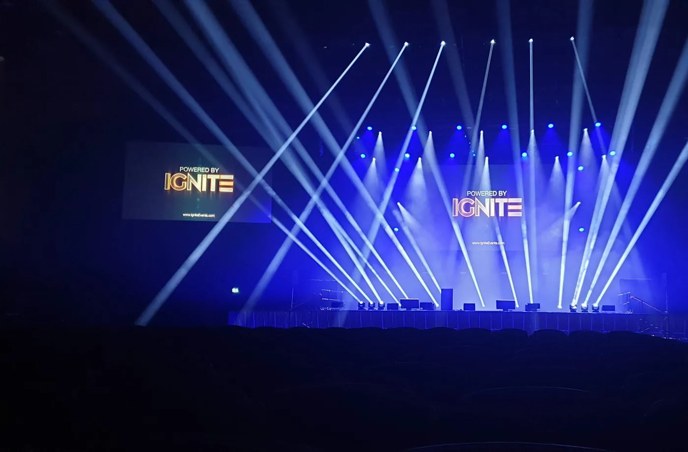 Arena stage with 'Powered by IGNITE' on the screens and white beam lighting fanning over empty seats