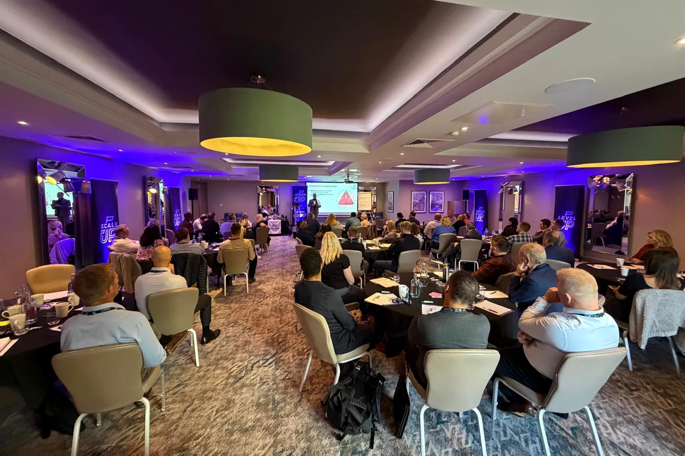 Seminar in a hotel conference suite: delegates at round tables facing a speaker and projection screen, with branded banners and purple uplighting